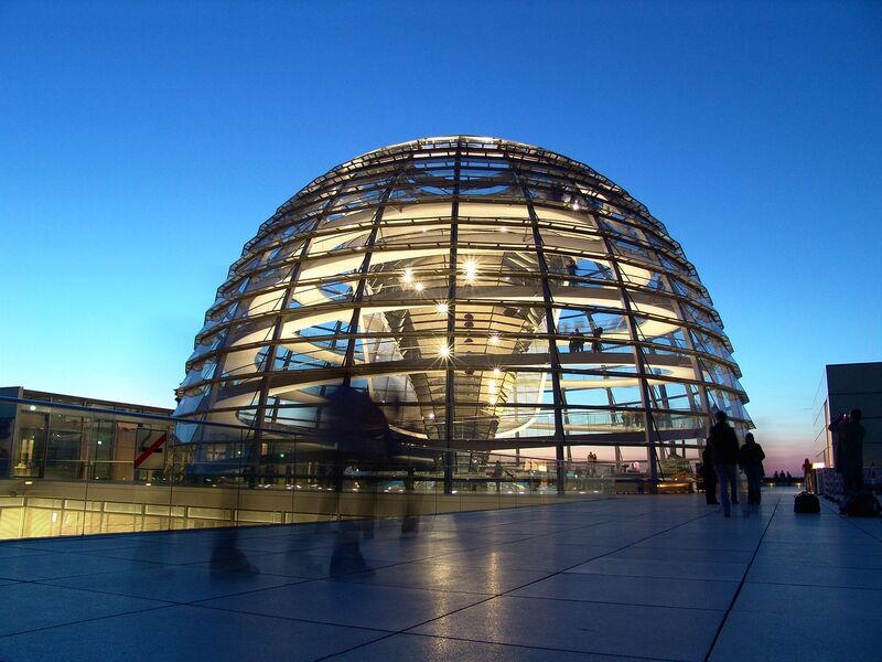Die beleuchtete Glaskuppel des Reichstagsgebäudes in Berlin leuchtet abends, während Menschen die Terrasse erkunden.