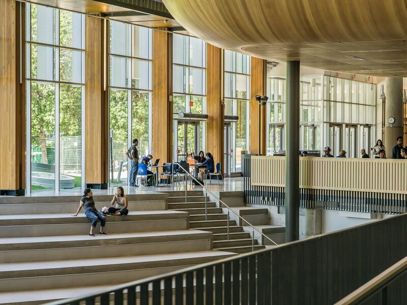 Blick ins Foyer einer Universität mit Studierenden, die auf Treppenstufen sitzen und sich unterhalten.
